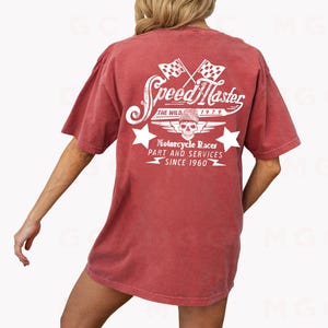 May include: A red t-shirt with a white graphic design on the back. The graphic features a skull with wings, two checkered flags, and the text "Speed Master The Wild 1979 Motorcycle Races Part and Services Since 1960".