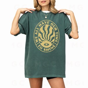 May include: A green t-shirt with a yellow circle graphic that says "We all have our time to shine" with a stylized eye and sunburst design.