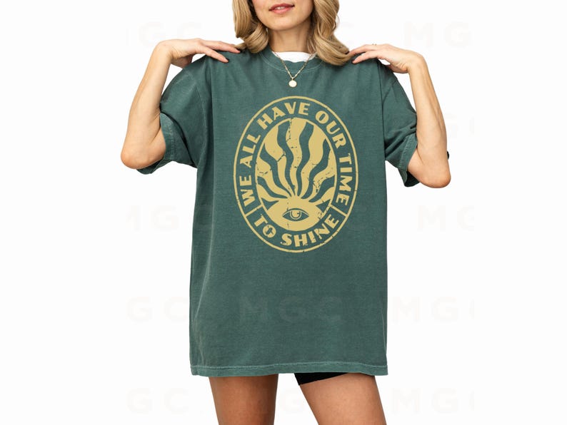 May include: A green t-shirt with a yellow circle graphic that says "We all have our time to shine" with a stylized eye and sunburst design.