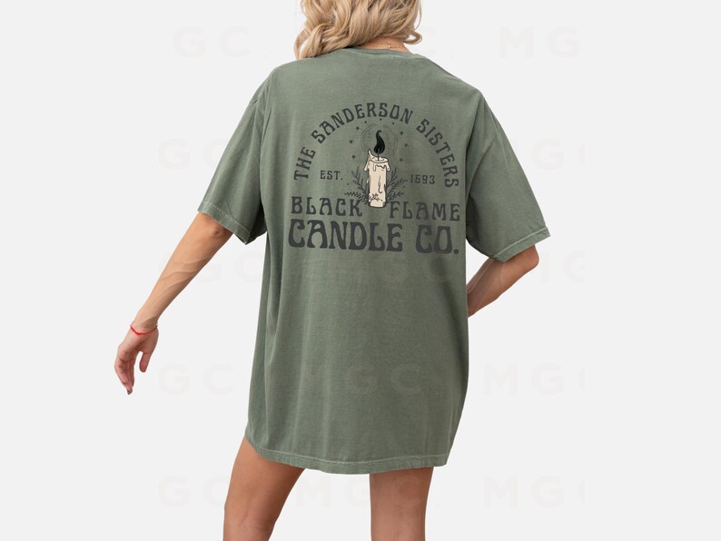 Pu&ograve; includere: Una maglietta verde con una stampa grafica nera sul retro. La grafica presenta una candela con una fiamma e il testo "The Sanderson Sisters Est. 1693 Black Flame Candle Co."