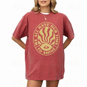 May include: A red t-shirt with a vintage-style graphic print that reads "We all have our time to shine". The graphic features a sunburst design with an eye in the center.