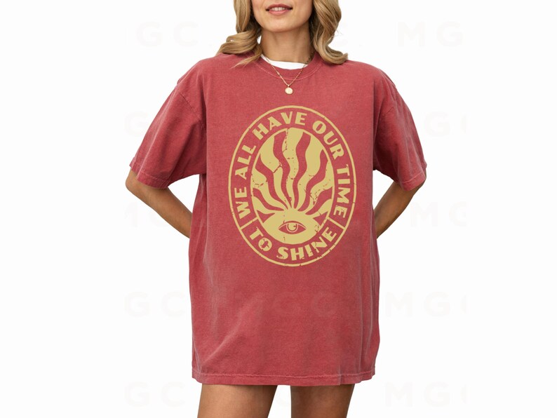 May include: A red t-shirt with a vintage-style graphic print that reads "We all have our time to shine". The graphic features a sunburst design with an eye in the center.
