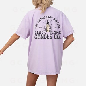 Pu&ograve; includere: Una maglietta color lavanda con una stampa grafica di una candela e il testo "The Sanderson Sisters Est. 1693 Black Flame Candle Co."