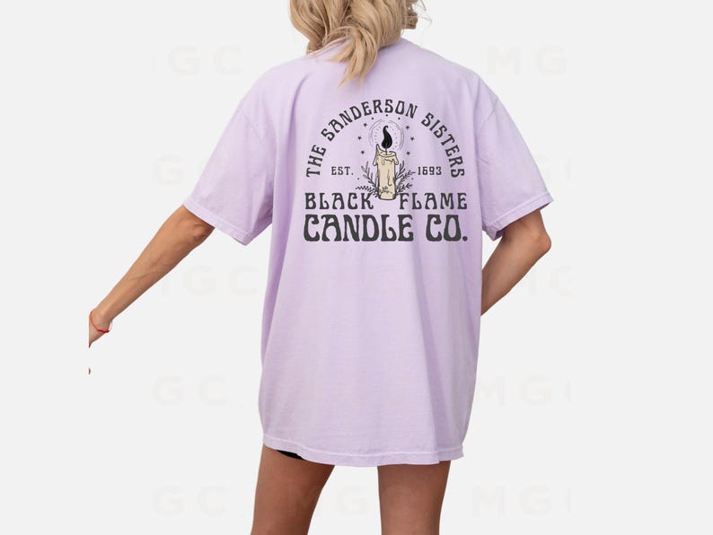 Pu&ograve; includere: Una maglietta color lavanda con una stampa grafica di una candela e il testo "The Sanderson Sisters Est. 1693 Black Flame Candle Co."
