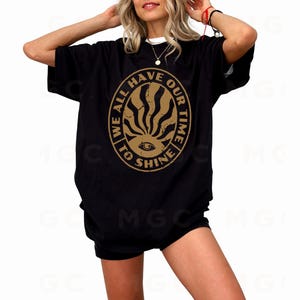 May include: Black oversized t-shirt with a gold graphic print of a circle with the text "We all have our time to shine" and a stylized eye with sun rays.