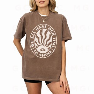 May include: A brown t-shirt with a white graphic print of a sun with an eye in the center. The graphic has the text "We all have our time to shine" around it.