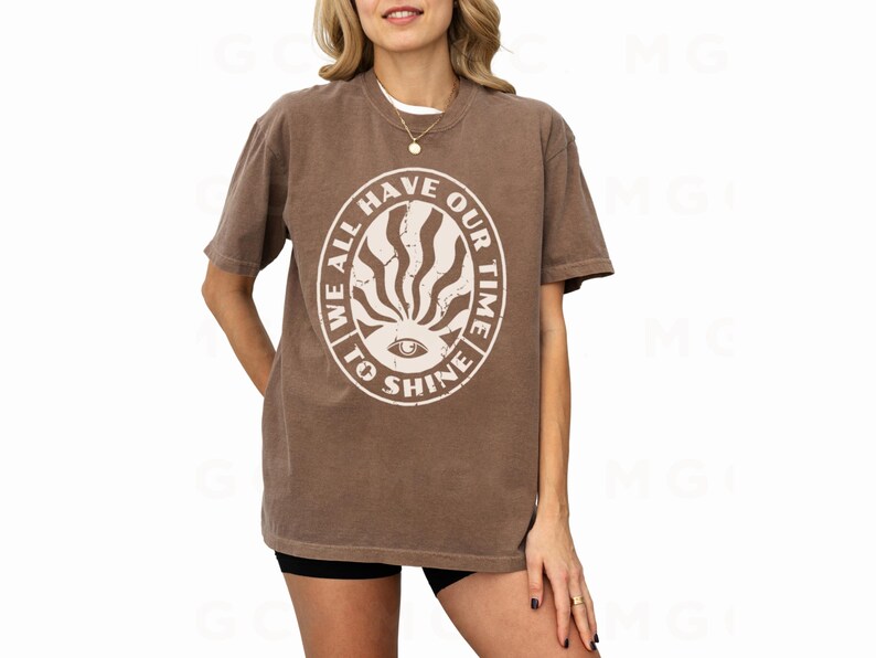 May include: A brown t-shirt with a white graphic print of a sun with an eye in the center. The graphic has the text "We all have our time to shine" around it.