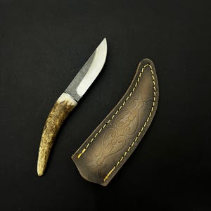 May include: A small, handcrafted knife with a polished steel blade and a handle made from antler. The knife is accompanied by a brown leather sheath with decorative stitching and embossed design.