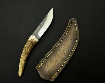 Puede incluir: Un pequeño cuchillo artesanal con una hoja de acero pulido y un mango de asta. El cuchillo se presenta con una funda de cuero marrón con costuras decorativas y un diseño en relieve.