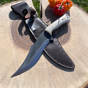 May include: A black-handled knife with a curved blade and a leather sheath. The knife is resting on a wooden surface with three red and orange peppers.