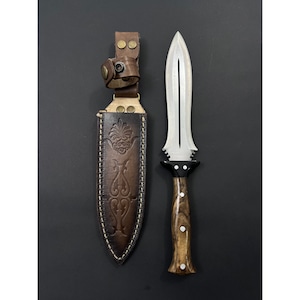 Könnte beinhalten: Eine braune Lederscheide mit einem dekorativen Design und ein silberner Dolch mit einem Holzgriff.