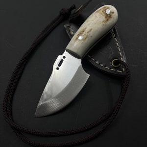 Puede incluir: Un cuchillo pequeño con mango blanco, hoja curva y funda de cuero. El cuchillo está sujeto a un cordón marrón.