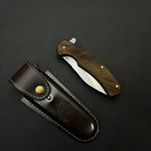 May include: A folding pocket knife with a wooden handle and a stainless steel blade, next to a brown leather sheath. The sheath has a snap closure and a decorative embossed design. The knife is approximately 8 inches long when open.