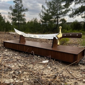 May include: A silver decorative sword with Arabic calligraphy on the blade, displayed on a wooden stand. The sword has a gold-colored hilt and a brown wooden handle.