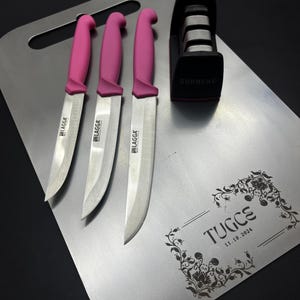 May include: A silver metal cutting board with a floral design and the text "TUGCS 11.19.2024". Three pink handled knives and a red and black knife sharpener are on the board.