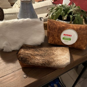 May include: Three different fabric cosmetic bags. One is white faux fur, one is brown and white cowhide, and the other is brown leather with a Huggies Natural Care baby wipe container visible inside.