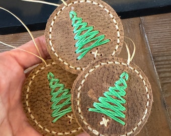 May include: Three brown leather ornaments with green stitched Christmas trees. Each ornament has a white stitched border and a loop for hanging.