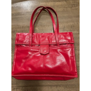 Puede incluir: Un bolso tote de cuero rojo brillante con dos asas. El bolso tiene forma rectangular con cierre de solapa y un bolsillo delantero. El bolso está hecho de cuero liso y brillante.