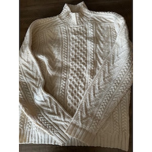 May include: An off-white long-sleeve sweater featuring a high neck with a short zipper closure. The garment is crafted with a complex cable knit pattern, including vertical columns of braided textures and honeycomb stitches across the front.