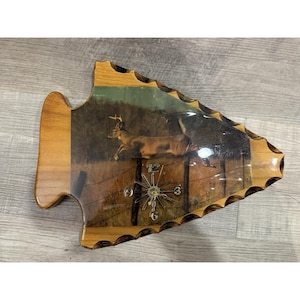 Könnte beinhalten: Eine pfeilspitzenförmige Uhr aus Holz mit einer Wildtierszene, die einen über einen Zaun springenden Hirsch zeigt. Die Uhr hat ein goldfarbenes Zifferblatt und Zeiger. Die Uhr ist aus Holz mit einer transparenten Schutzbeschichtung.