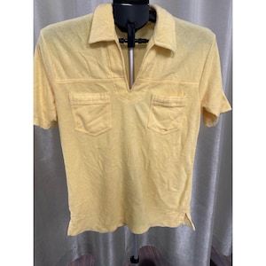 Peut inclure: Un polo jaune à manches courtes avec un col en V et deux poches avant. La chemise est faite d'un tissu doux et texturé et a une coupe décontractée. La chemise est présentée sur un cintre noir.