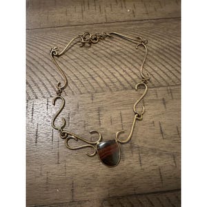 May include: A handcrafted necklace with a bronze-colored metal chain and a central, shield-shaped stone. The stone features a mix of brown, red, and black hues. The chain has a unique, curved design.