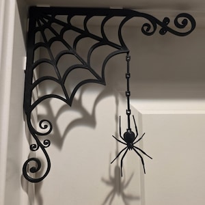 May include: A black corner bracket featuring a spider web design with decorative scrollwork at the ends. A small black spider hangs from the web by a thin chain. The bracket is mounted in the upper corner of a door frame, casting a shadow.