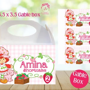 May include: White gable box with a handle, featuring a pink and white checkered pattern. The box is decorated with a cartoon strawberry girl, the name "Amina Shortcake", and the number 2. The box measures 6 x 3.5 x 3.5 inches.