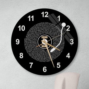 Puede incluir: Reloj de pared redondo negro con manecillas doradas y números blancos. La esfera del reloj tiene un diseño de disco de vinilo con las palabras "SONG NAME" en el centro. Una escultura de rostro abstracto blanco está debajo.