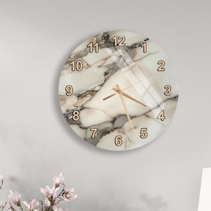 Può includere: Un orologio da parete rotondo con un design marmorizzato. Il quadrante presenta un motivo marmorizzato bianco e grigio con numeri e lancette dorate. L'orologio è appeso a una parete grigio chiaro.