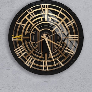 May include: A round black wall clock with gold-colored Roman numerals and hands. The clock face features a geometric design with a maze-like pattern. The clock is mounted on a light gray wall.