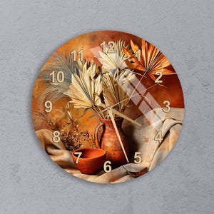 Puede incluir: Reloj de pared redondo con un diseño de naturaleza muerta. Presenta un jarrón y un cuenco de terracota, plantas secas y una tela de tonos neutros. La esfera del reloj tiene una paleta de colores cálidos con una superposición de vidrio y números negros.