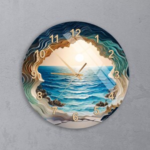 May include: A round wall clock featuring a layered, topographic-style border in shades of blue, teal, and tan that frames a central ocean scene. The clock face displays gold-colored numbers from 1 to 12 and matching gold-colored clock hands.