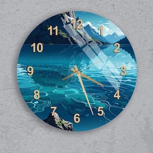 Op de afbeelding: Ronde wandklok met een landschapsontwerp met een meer en bergen. De wijzerplaat toont een blauw-turkooise waterpartij met goudkleurige wijzers en cijfers. De klok is aan een grijze muur bevestigd.