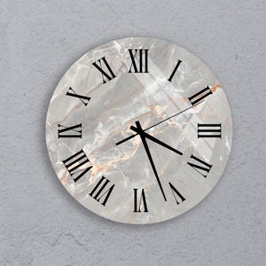 May include: Round wall clock with a marble-like face featuring black Roman numerals and hands. The clock has a gray and gold color scheme, with a glossy finish. The clock is mounted on a gray wall.