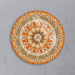 May include: Round wall clock with a detailed mandala design in shades of orange, brown, and cream. The clock face features a radial pattern with a central focal point and traditional numerals. The clock is mounted on a gray wall.