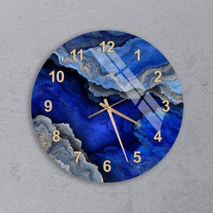 May include: Round wall clock with a blue and gold agate design. The clock face features gold numerals and hands, set against a swirling blue background. The clock is mounted on a gray wall.