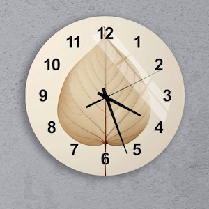 Peut inclure: Horloge murale ronde avec un cadran crème et des chiffres noirs. L'horloge présente un motif de feuille marron clair détaillé au centre. Les aiguilles noires des heures et des minutes indiquent l'heure.