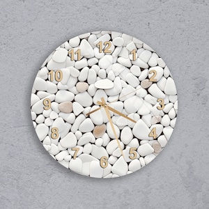 May include: Round wall clock with a white pebble design. The clock face features gold-colored numerals and hands. The clock's design incorporates a mix of white and tan-colored pebbles, creating a textured appearance. The clock is mounted on a gray wall.