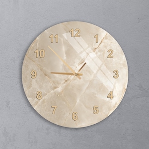 May include: A round wall clock featuring a beige marble-patterned face with gold-colored numbers from 1 to 12. The clock has three thin gold-colored hands and a smooth, reflective surface finish that displays subtle light reflections.