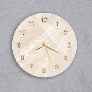 May include: Round wall clock with a marble-like face in cream and beige tones. The clock features gold-colored hands and numerals. The numerals are in a simple, elegant font. The clock is mounted on a gray wall.