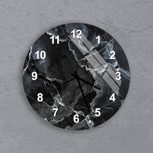Puede incluir: Reloj de pared redondo con diseño de mármol negro. La esfera del reloj presenta números blancos y manecillas negras. El reloj está montado en una pared gris.