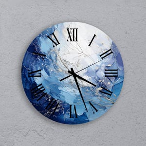 Puede incluir: Un reloj de pared redondo con un diseño abstracto azul, blanco y dorado. La esfera del reloj presenta marcadores de hora en números romanos y manecillas negras.