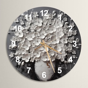 Puede incluir: Un reloj de pared redondo con fondo gris y flores blancas en 3D. La esfera del reloj tiene números negros del 1 al 12 y manecillas doradas.