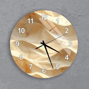 May include: Circular wall clock featuring a desert scene. The clock face has a beige and tan colour scheme, depicting sand dunes. Black clock hands and white numerals are present. The clock is mounted on a grey wall.