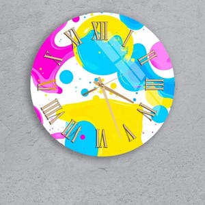 May include: Round wall clock with a colorful abstract design. The clock face features a white background with splashes of yellow, blue, and pink. Gold-colored Roman numerals mark the hours, and gold hands indicate the time.