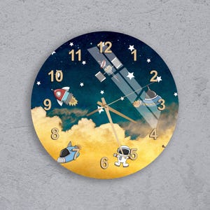 Könnte beinhalten: Runde Wanduhr mit Weltraum-Thema. Das Zifferblatt hat einen blau-gelben Hintergrund mit Illustrationen einer Rakete, eines Raumschiffs, eines Astronauten und Sternen. Die Zeiger der Uhr sind goldfarben.
