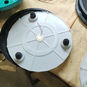 Op de afbeelding: Een close-up van een witte draaitafel met vier zwarte rubberen voetjes en een kleine zilveren sensor in het midden. De draaitafel is omgeven door draden en andere componenten.