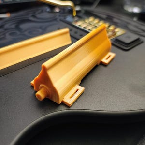 Może przedstawiać: Trójwymiarowe, drukowane, pomarańczowo-żółte elementy z tworzywa sztucznego. Jeden element to prostokątny pręt, a drugi ma złożony kształt z cylindrycznym występem i prostokątnymi wycięciami. Obiekty znajdują się na czarnej powierzchni.