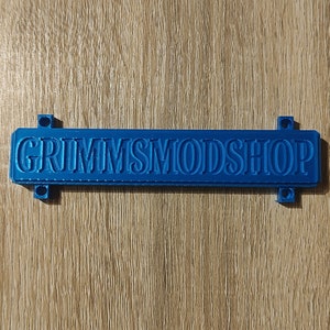 Puede incluir: Un objeto rectangular azul con el texto "GRIMMSMODSHOP" está centrado en una superficie de madera. El objeto tiene una fuente en relieve y pequeñas pestañas en cada extremo.